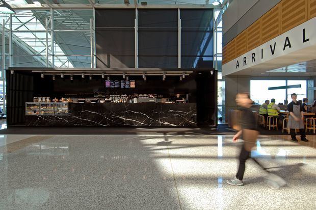 A spacious modern airport arrival hall featuring a dark café or food-service counter with dramatic veined stone cladding, polished terrazzo flooring, exposed steel roof structure, extensive glazing, and a prominent ARRIVAL sign. People and café seating occupy the right side of the terminal.