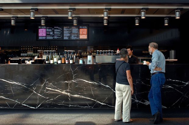 A dark, sophisticated commercial bar or café interior viewed from the customer area. A long black marble-clad service counter with dramatic white veining dominates the foreground, backed by shelves of bottles, coffee or bar equipment, and a black menu wall. Warm timber ceiling battens and numerous directional spotlights provide contrast, while staff and customers stand at the counter.