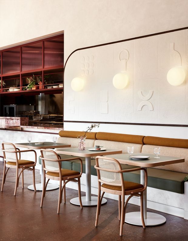A refined contemporary restaurant or hospitality dining interior with closely arranged two-person tables, curved cane-back timber chairs, and a long upholstered banquette in ochre mustard. A dark reddish-brown service counter and open shelving occupy the rear, while a large white textured feature wall with embossed abstract motifs and glowing spherical wall lights forms the main backdrop. The space has polished warm brown flooring and a restrained palette of cream, timber, mustard, and deep burgundy.