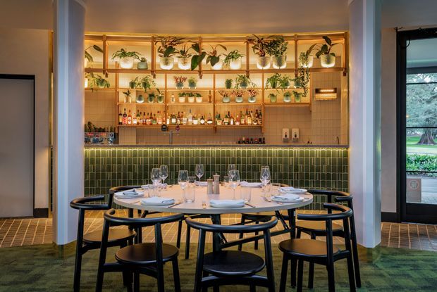 A warmly lit restaurant dining area with a round communal table surrounded by black timber chairs. Behind it is a tiled bar with a green glazed-tile front, open shelving filled with bottles and glassware, and abundant potted plants displayed on illuminated timber shelves. The space has a refined botanical atmosphere with dark green and terracotta-toned finishes.
