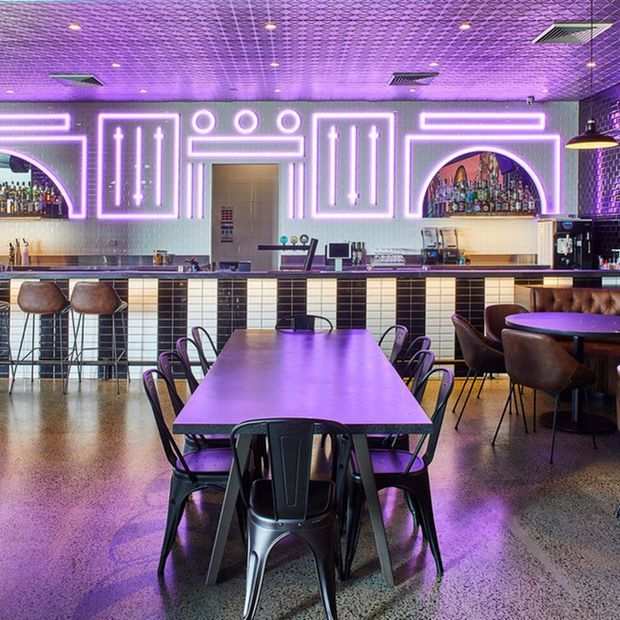 A vibrant contemporary bar and dining venue with a long central communal table, black metal chairs, leather booth seating, a tiled bar front, and a back bar illuminated by vivid purple and white neon geometric detailing. The space has a polished concrete floor, exposed dark ceiling grid, pendant lighting, and a lively nightlife atmosphere.