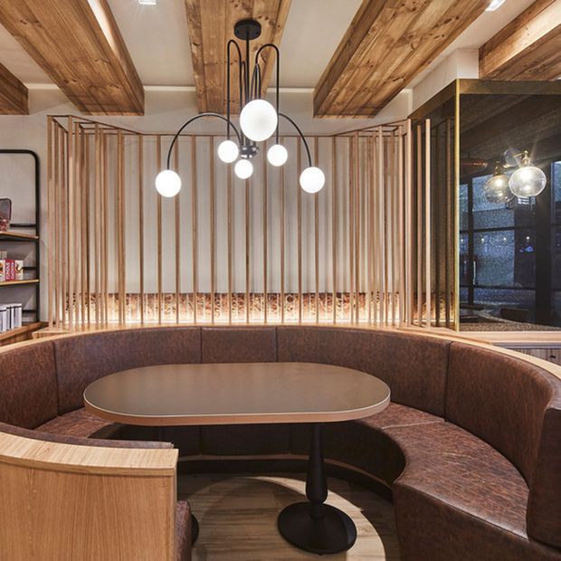 A warm, refined built-in dining nook with a large curved upholstered banquette surrounding an oval table. Vertical timber slat screening, exposed timber ceiling beams, timber flooring, dark-framed glazing, and a sculptural cluster of spherical pendant lights define the space.