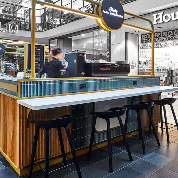 A contemporary coffee bar kiosk in a shopping centre, featuring a long white counter, dark timber vertical panelling, teal ribbed tile detailing, brass-coloured framing and trim, black bar stools, and commercial coffee equipment. Large glazed shopfronts and visible signage surround the open café counter.