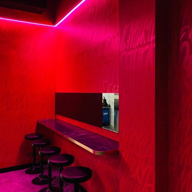 A compact, intensely red entertainment or bar-like interior with textured red walls, vivid pink LED strip lighting along the ceiling edges, a narrow wall-mounted counter, a dark mirror or reflective panel, and several black stools. The atmosphere is dramatic and nightclub-inspired.