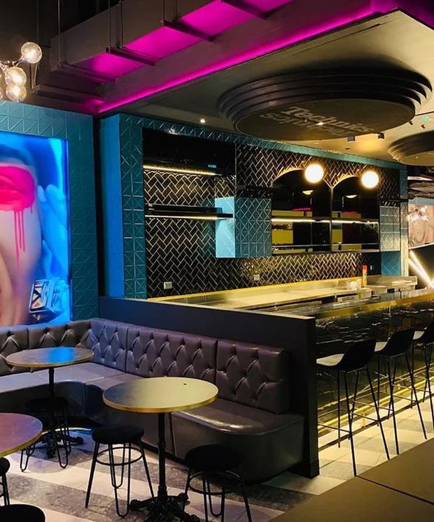 A vibrant, upscale commercial bar and lounge interior with a long marble-look bar, black herringbone tile backsplash, illuminated shelving, tufted dark banquette seating, round café tables, and high stools. The space features bold cyan blue wall tiles, dramatic magenta cove lighting, globe pendant lights, and a large colorful mural, creating a theatrical nightlife atmosphere.