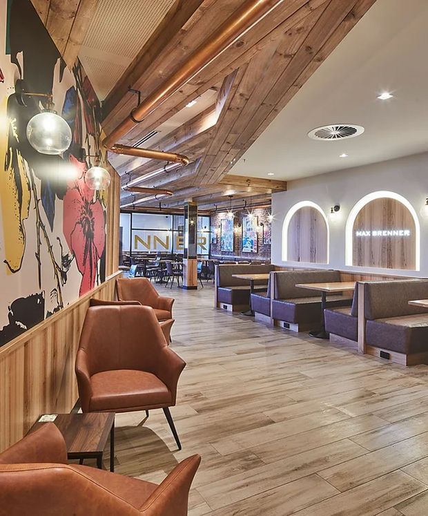 Interior view of a contemporary chocolate café or restaurant, featuring booth seating, leather lounge chairs, timber-look flooring, extensive timber ceiling panelling, exposed copper services, arched illuminated wall niches, and bold mural artwork. The space has a warm, industrial-inspired hospitality fit-out with open dining and seating areas.