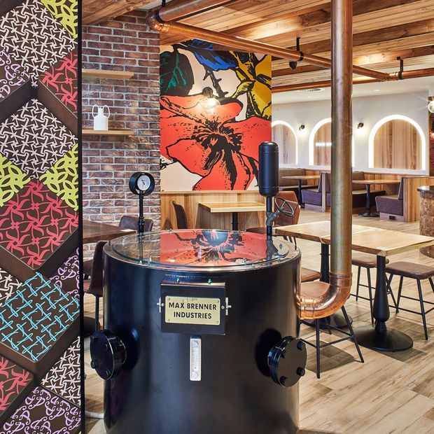 A vibrant industrial-style chocolate restaurant interior featuring a large black chocolate-making vessel in the foreground, exposed copper piping, brick walls, timber ceiling boards, patterned graphic wall panels, bold pop-art artwork, arched booth openings, and timber dining tables with upholstered seating.