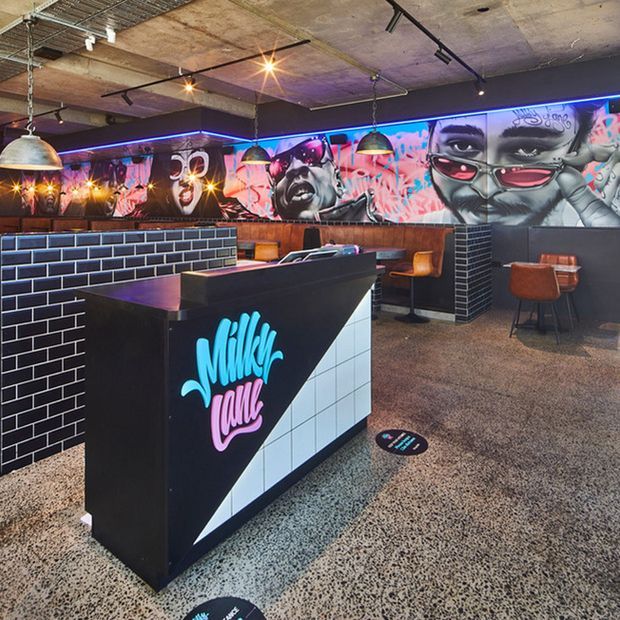 A vibrant urban-style milkshake bar or café interior with a black tiled service counter, illuminated neon branding, terrazzo flooring, exposed concrete ceilings, track lighting, timber wall panelling, and colourful graffiti-style mural artwork. Bar seating lines the rear wall, creating a bold, playful hospitality space.