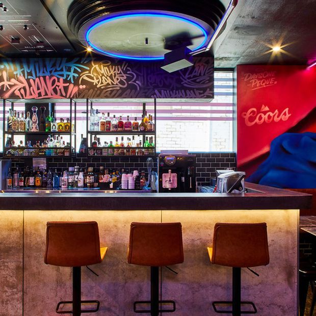 A vibrant contemporary bar interior with a three-seat counter, open shelving stocked with bottles and glassware, dark subway-tile backsplash, exposed concrete ceiling, graffiti-style artwork, and colorful neon accent lighting. The bar front has a warm illuminated finish, while the stools are upholstered in brown leather.