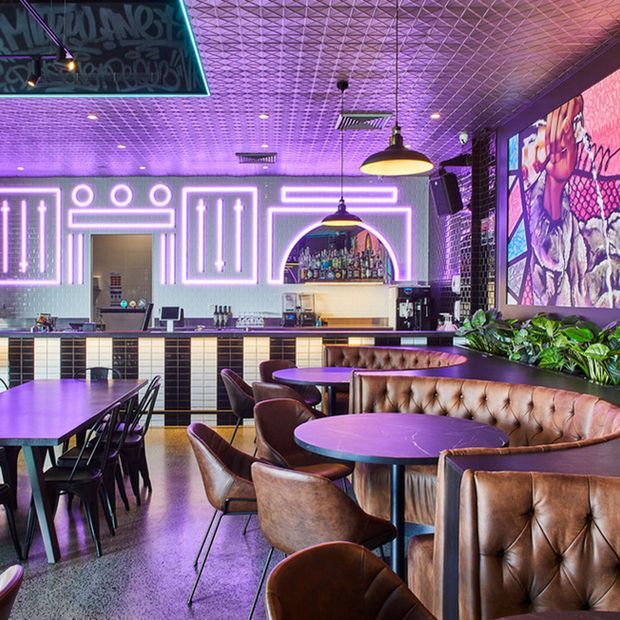 Vibrant contemporary bar and dining venue with tufted leather booth seating, round tables, bar stools, a tiled bar counter, illuminated geometric wall detailing, pendant lights, patterned ceiling, large artwork, and purple neon-style ambient lighting.