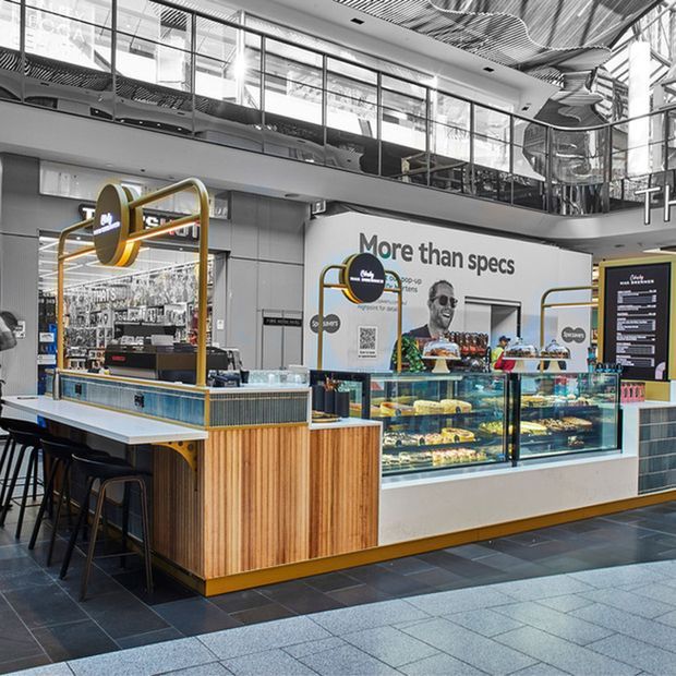 A contemporary café or food-service kiosk in a large commercial interior, featuring timber-fronted counters, glass pastry display cases, dark tiled flooring, brass-gold framing and signage, and a prominent mezzanine with extensive glazing and reflective metal detailing.