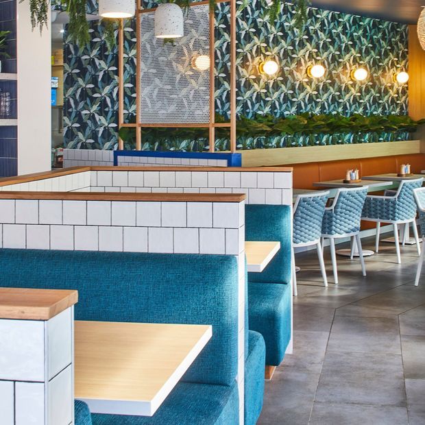 A vibrant contemporary commercial dining interior, likely a café or restaurant, featuring turquoise upholstered booth seating, light timber tables, white rectangular tiled partitions with timber caps, patterned botanical wall covering, integrated greenery, decorative pendant lights, and a dining area with matching blue chairs. The space has a lively, fresh, highly coordinated fit-out.