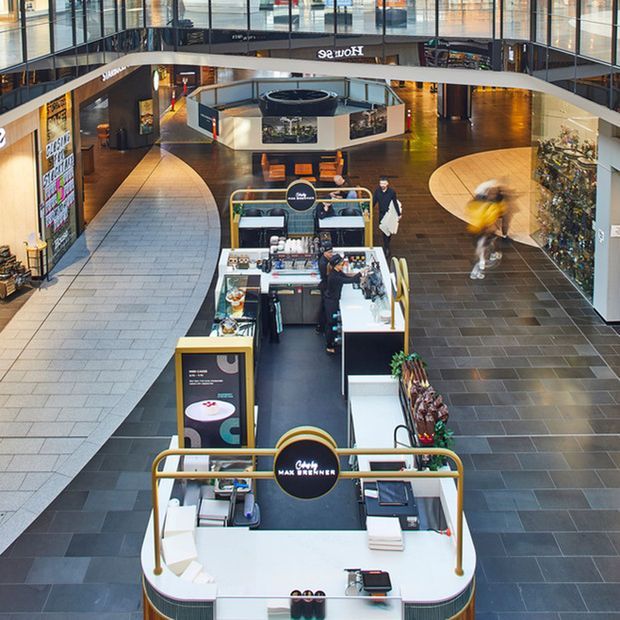 A contemporary multi-level shopping-centre atrium viewed from above, with curved balconies, large glazed shopfronts, dark tiled flooring, and several branded coffee kiosks arranged along the central circulation space. Shoppers and retail displays are visible throughout.
