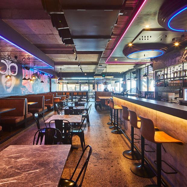 Interior view of a lively modern restaurant or bar, with booth seating, marble-top dining tables, a long illuminated bar counter, bar stools, exposed concrete ceilings, visible services, large graphic wall art, and vivid blue and pink linear and circular LED lighting.