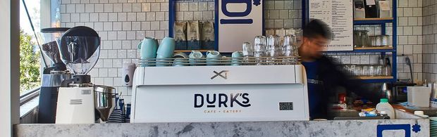 A compact contemporary café counter with a white commercial espresso machine, grinder, coffee equipment, cups, glassware and turquoise ceramic vessels. The backdrop features white subway-tile walls, dark blue shelving and a grey stone-look counter, creating a clean, casual service area.