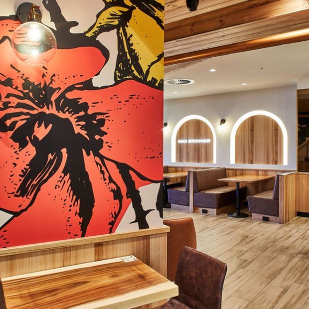 A warmly lit commercial dining space with booth seating, timber tables and wall panelling, illuminated arched wall niches, and a bold oversized black, coral-red and yellow graphic mural. The interior combines natural timber finishes, upholstered seating, pendant lighting and a polished casual restaurant fit-out.