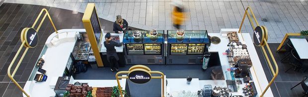 Overhead view of a contemporary retail food-and-beverage kiosk or café concession in a tiled commercial setting. The counter features dark display cases, refrigerated food displays, confectionery and packaged goods, with distinctive yellow structural rails and signage. Customers and staff are visible around the compact service area.