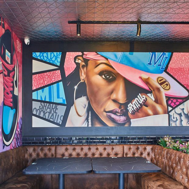 A vibrant commercial hospitality interior featuring a tufted brown leather banquette wrapping around dark tables, a large colourful hip-hop-inspired mural, patterned geometric ceiling panels, and linear black track lighting. The space has a bold, urban, artistic atmosphere.