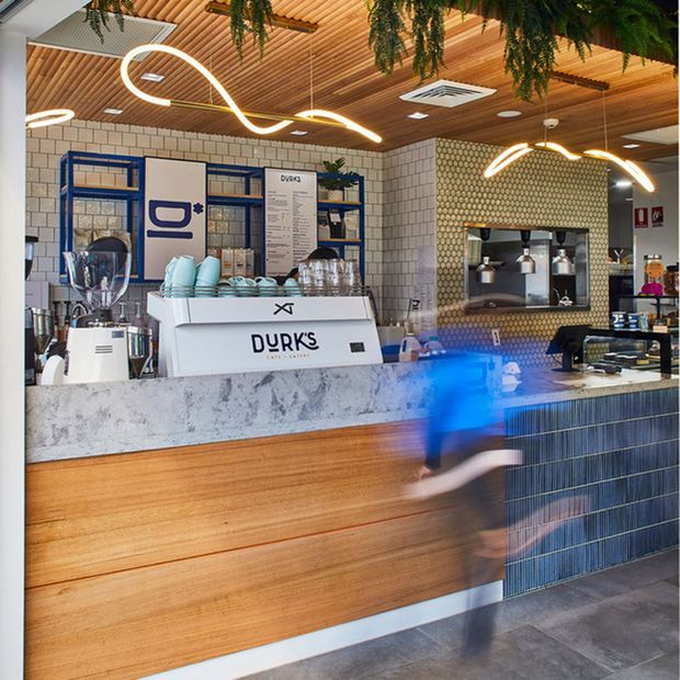 A contemporary café interior focused on a long service counter. The counter combines warm horizontal timber panelling with a mottled grey stone-look benchtop and a dark blue vertically tiled front section. Behind it are blue-framed open shelving units, coffee equipment, a menu board, and a recessed coffee station set into a patterned light wall tile. The ceiling features extensive timber slat lining, recessed services, hanging greenery, and sculptural linear pendant lighting. A blurred person is moving in the foreground.