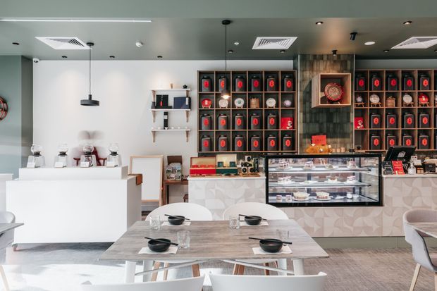 Bright contemporary café interior with a white service counter, refrigerated pastry display, dining tables, and extensive dark built-in shelving filled with colourful packaged goods and decorative objects. The space features a patterned tiled counter front, timber-look wall cladding, pendant lighting, recessed ceiling lights, and a clean, modern retail dining atmosphere.