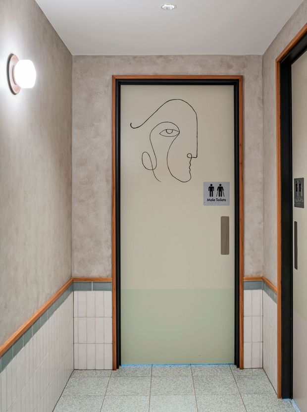 A narrow commercial hallway leading to a male toilet entrance. The corridor has textured grey plaster walls, white tiled lower walls with teal trim and timber edging, terrazzo-style floor tiles, dark-framed doors with warm timber surrounds, and a frosted glass toilet door featuring minimalist line-art signage. A round wall light and recessed ceiling light provide illumination.