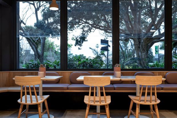 A warm, contemporary dining or café-style seating area arranged along a built-in banquette beneath large black-framed windows. Light timber spindle-back chairs face a long timber counter, with pendant lighting and small table lamps. Dense greenery and mature trees are visible outside, while dark wall finishes and rich brown upholstery create a refined, inviting interior.
