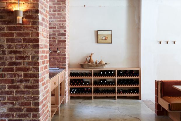 A rustic wine storage area set within a brick-lined interior. Low open timber wine racks hold numerous bottles against a white plaster wall, with a concrete floor, exposed red brickwork, a wall sconce, and a partial built-in leather-upholstered bench or dining nook visible at right.