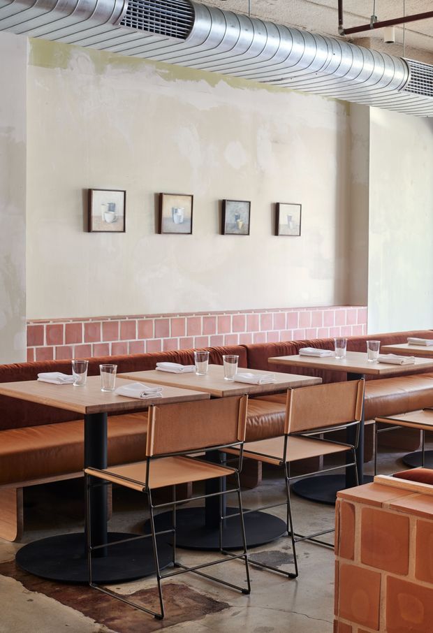 A warmly styled restaurant dining area with long banquette seating, timber tables, leather-upholstered benches, and slim black metal-framed chairs. The space features an exposed silver HVAC duct and ceiling services, a lightly textured plaster wall, a pink-red glazed brick dado, framed artworks, and a polished concrete floor. The composition has a contemporary hospitality character with industrial architectural detailing and warm earthy furnishings.
