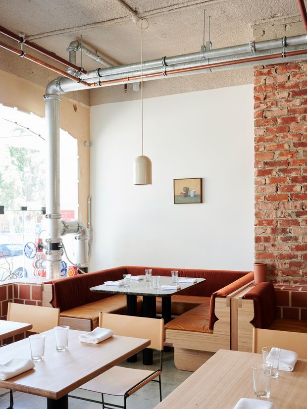 Interior view of a bright restaurant dining area with exposed industrial services, including suspended metal ductwork and red-painted pipes across a textured ceiling. The space features a red exposed-brick wall, pale plaster walls, timber dining tables, black metal table bases, and built-in rust-coloured upholstered banquette seating. Large glazing brings in daylight, while simple pendant lighting and minimal wall art complete the casual contemporary-industrial setting.