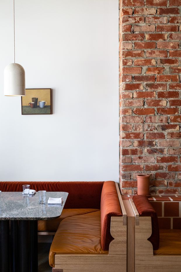 A stylish dining nook with a built-in rust-orange leather banquette surrounding a grey stone-topped table. The space combines a white plaster wall with an exposed red brick feature wall, a suspended cream pendant light, timber detailing, and a small framed artwork. The composition has a warm, refined restaurant or hospitality-inspired character with strong mid-century and industrial influences.