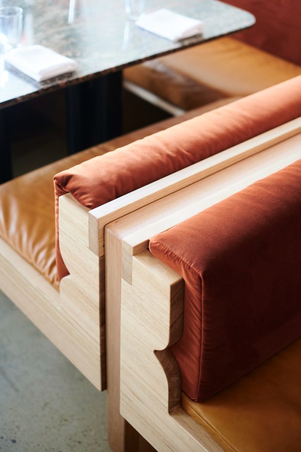 Close-up interior detail of custom timber-framed upholstered seating in warm rust-brown fabric, positioned beside a dark stone-topped table or counter. The pale timber joinery has sculptural stepped detailing, with a polished concrete floor visible below.