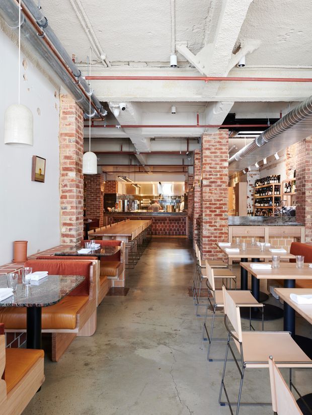 Interior view of a large industrial-style restaurant or café with exposed painted concrete ceiling, visible ductwork and red fire-suppression pipes, exposed red brick columns and walls, polished concrete flooring, booth seating, dining tables, pendant lights, and a bar with extensive bottle storage at the rear.