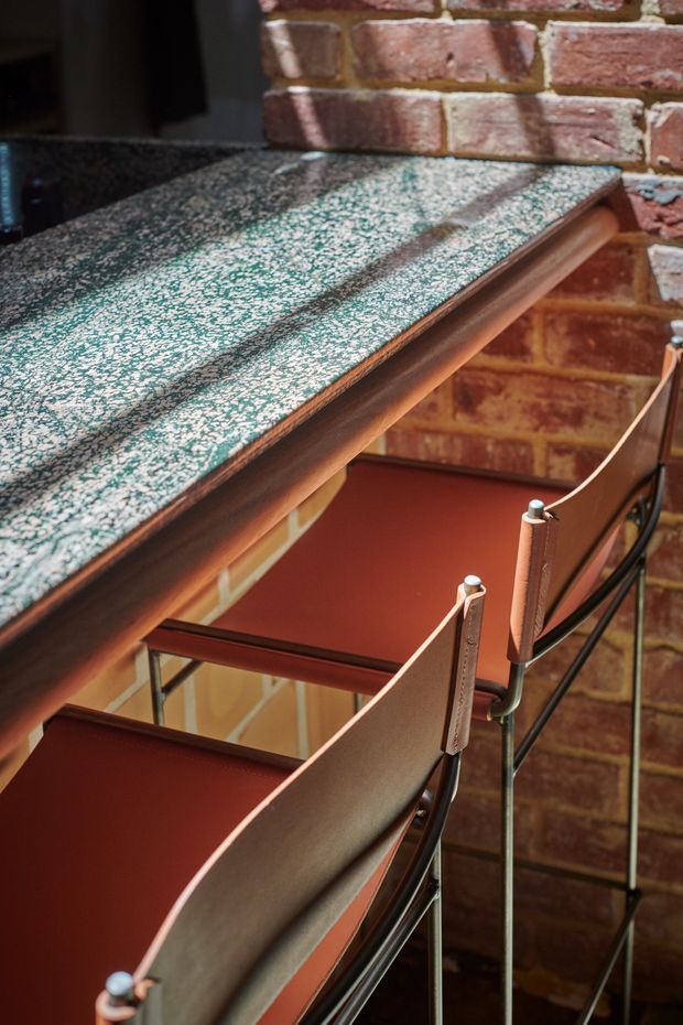 Close-up architectural detail of a narrow stairway or landing beside an exposed red-brick wall. The stairs feature dark metal railings, warm brown leather or upholstered balustrade panels, and a richly textured green-and-white stone or terrazzo-like ledge. The composition highlights industrial materials and crafted detailing in a warm, moody setting.