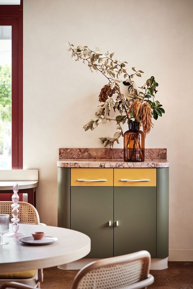 A styled dining area featuring a compact mid-century-inspired sideboard with muted olive-green doors, mustard-yellow drawers, and a polished reddish-brown stone top. A large amber glass vase with dried branches sits on the sideboard against a warm beige wall. In the foreground is a round light-colored dining table with woven cane chairs, while a deep red window and trim are visible at left.