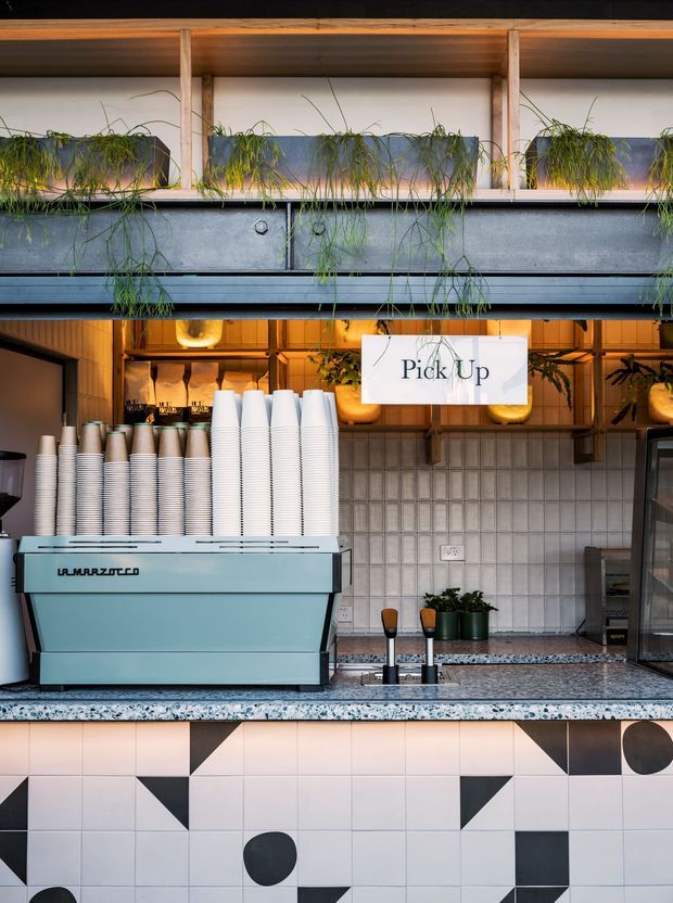 A contemporary café coffee pickup counter featuring a pale blue La Marzocco espresso machine, stacks of takeaway cups, a terrazzo countertop, geometric patterned tiled cabinetry, warm timber shelving, hanging plants, and a prominent “Pick Up” sign. The space has a carefully styled, modern hospitality interior with warm ambient lighting and strong geometric detailing.