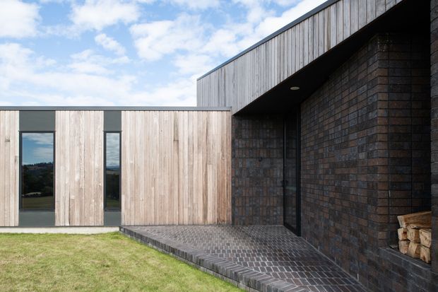 Exterior view of a low-profile contemporary home with a sharply projecting roof volume, vertical weatherboard cladding, dark brick walls, narrow vertical glazing, and a paved entry terrace. The building forms a sheltered courtyard-like approach, with lawn in the foreground and a partly cloudy sky beyond.