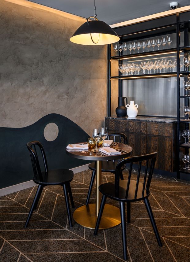 A moody, refined restaurant dining area with a small round table set for two, black bentwood-style chairs, a sculptural black and gold pendant light, textured grey plaster walls, and a dark timber bar or display cabinet stocked with wine glasses. The floor has a dark patterned finish with geometric light grout lines, creating a sophisticated hospitality interior.