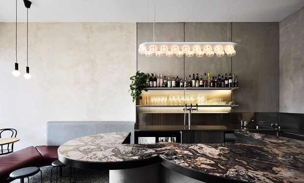 A sophisticated contemporary home bar and lounge featuring a large sculptural dark stone bar counter with a rounded marble-patterned extension, dark integrated cabinetry, open bottle and glassware shelving, a sink and tap, and a striking linear pendant light. A grey sofa and burgundy seating occupy the foreground, against textured grey wall panels and a pale plaster wall.
