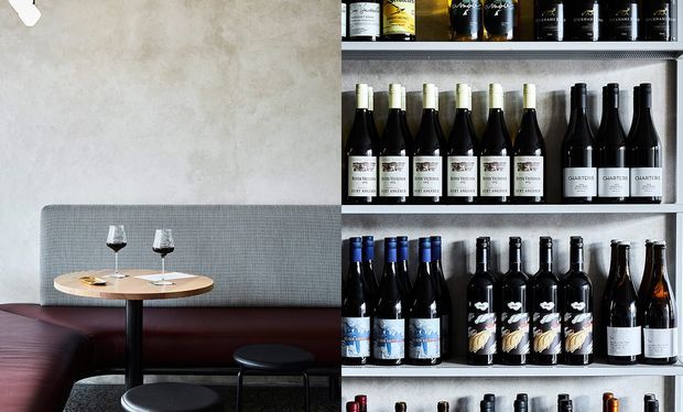 A stylish wine bar or tasting area combines a small round dining table with wine glasses, upholstered banquette seating and stools on the left, while a tall open shelving unit displaying rows of wine bottles dominates the right. The space has a refined, contemporary hospitality feel with textured grey walls and upholstery, dark accents, and warm timber.