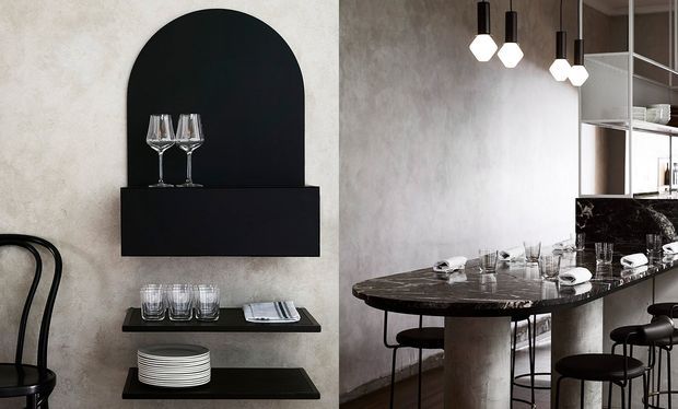 A sophisticated bar and dining interior featuring a dark marble-clad island with rounded ends, black stools, pendant lights, and an adjacent wall-mounted black shelving unit with an arched display niche holding glassware. The space has textured pale plaster walls, dark finishes, and refined contemporary detailing.