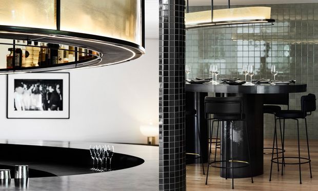 A sophisticated contemporary dining and entertaining space divided by a glossy black tiled column. On the left is a curved black kitchen or bar counter beneath a large sculptural circular pendant light, with a framed monochrome artwork on the white wall. On the right, a dark dining table and bar-height seating sit against a wall of small pale green-grey square tiles, with a linear pendant light overhead. The room combines black glossy surfaces, metallic details, pale tiled walls, and light timber flooring.