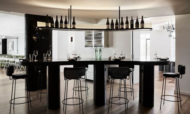 A dramatic monochrome home bar or entertaining space with a long glossy black counter, numerous black bar stools, suspended shelving displaying wine bottles, and glassware-lined cabinetry behind. White walls and ceiling contrast with black tiled and joinery elements, while timber flooring adds warmth. Dining or kitchen areas are visible in the background.