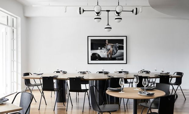 Bright, minimalist dining room with a long communal dining table set for meals, multiple black chairs, a smaller round timber table in the foreground, pale timber flooring, white walls, large windows, and distinctive black-and-white pendant and track lighting. A framed artwork provides a focal point on the rear wall.