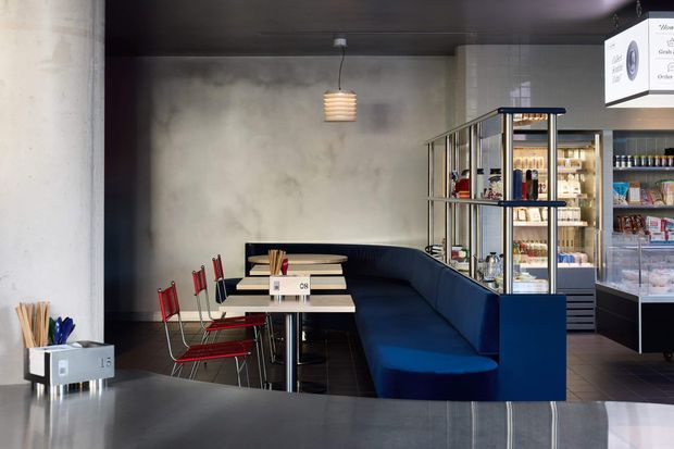A modern commercial café or casual dining space with a long built-in deep blue banquette, small rectangular tables, and red metal chairs. The room has a dark ceiling, raw mottled concrete-look walls, black-framed glazed shelving, pendant lighting, and a retail service/display area with packaged goods at the right. A dark counter edge is visible in the foreground.