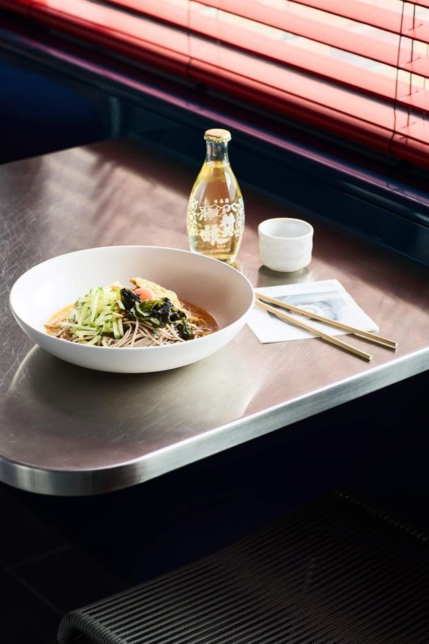 Close-up styled view of a dark, industrial-looking dining counter or table with a brushed stainless-steel surface and rounded edge. A bowl of noodles, a decorative bottle, ceramic cup, chopsticks, and papers are arranged on the tabletop. Red horizontal blinds and dark wall panelling appear in the background, with a dark woven chair or bench partially visible below.