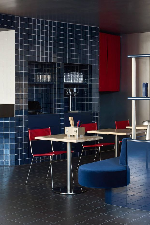A stylish compact commercial dining and bar interior with a deep navy blue tiled wall, integrated open shelving displaying glassware, dark tiled flooring, and a dark ceiling. Small square dining tables with polished metal pedestal bases are paired with vivid red chairs. A curved navy upholstered banquette wraps around the foreground, while red cabinetry and stainless-steel bar shelving add strong colour and material contrast. The space has a bold, refined, retro-modern hospitality aesthetic.