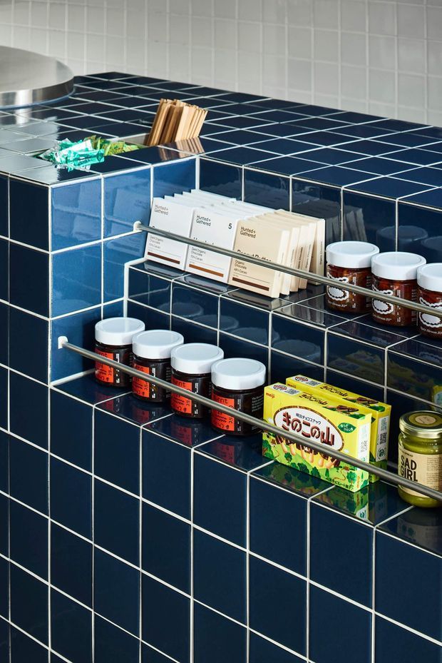 Close-up of a compact tiled pantry or kitchen storage area with glossy navy-blue square wall and counter tiles, white grout, integrated open shelving, and slim metal rails holding jars, tins, packaged goods, recipe cards, and utensils. A small portion of a stainless-steel fixture is visible at the upper left. The image has a bold, retro-inspired, highly graphic appearance with strong blue-and-white contrast.