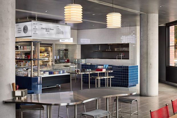 A modern commercial café or canteen interior with a compact food service counter, refrigerated display shelving, tiled service backsplash, dark blue tiled counter front, and a communal stainless-steel table with stools. The space features exposed concrete columns, a dark ceiling, graphic menu boards, pendant lights, and large black-framed windows.