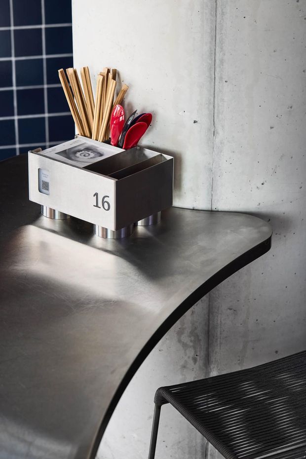 Close-up of a compact, industrial-style dining or hospitality setting. A dark stainless-steel or metal table with a curved edge holds a numbered metal cutlery and utensil container, with wooden chopsticks and red utensils. The backdrop combines raw concrete with a dark tiled wall, while a black woven chair is partially visible in the foreground.