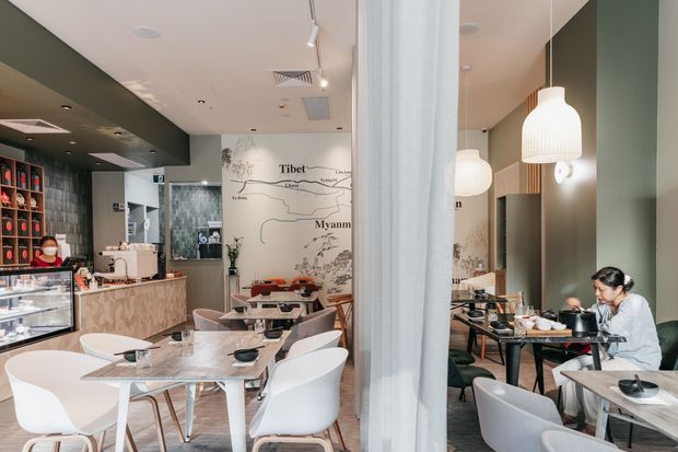 Interior view of a contemporary café or restaurant, showing a compact dining area with marble-topped tables, white molded chairs, pendant lights, a service counter, shelving, and a large illustrated wall mural. A curtain and mirror divide or reflect the dining space, with dark green walls and a polished commercial fit-out.